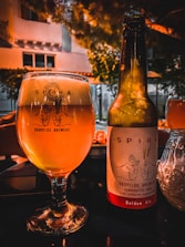A glass and a bottle of beer from Skopelos Brewers are placed on a table. The glass is filled with a golden-colored beer, and the bottle is labeled 'Golden Ale.' The background features a warm, orange-hued lighting with some greenery and parts of a building visible, suggesting an outdoor setting in the evening.