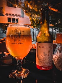 A glass and a bottle of beer from Skopelos Brewers are placed on a table. The glass is filled with a golden-colored beer, and the bottle is labeled 'Golden Ale.' The background features a warm, orange-hued lighting with some greenery and parts of a building visible, suggesting an outdoor setting in the evening.