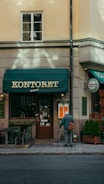 A street view of a building exterior with a storefront named 'Kontoret'. An older individual with a walking cane stands near the entrance, wearing a blue jacket and carrying a bag. The facade has a green awning and signs for a bar and restaurant. There is minimal foot traffic, and the building has a warm beige tone, contributing to a quiet, serene atmosphere.