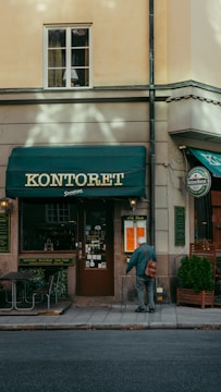 A street view of a building exterior with a storefront named 'Kontoret'. An older individual with a walking cane stands near the entrance, wearing a blue jacket and carrying a bag. The facade has a green awning and signs for a bar and restaurant. There is minimal foot traffic, and the building has a warm beige tone, contributing to a quiet, serene atmosphere.