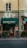 A street view of a building exterior with a storefront named 'Kontoret'. An older individual with a walking cane stands near the entrance, wearing a blue jacket and carrying a bag. The facade has a green awning and signs for a bar and restaurant. There is minimal foot traffic, and the building has a warm beige tone, contributing to a quiet, serene atmosphere.