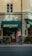 A street view of a building exterior with a storefront named 'Kontoret'. An older individual with a walking cane stands near the entrance, wearing a blue jacket and carrying a bag. The facade has a green awning and signs for a bar and restaurant. There is minimal foot traffic, and the building has a warm beige tone, contributing to a quiet, serene atmosphere.