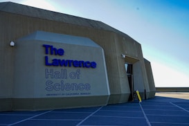 A large geometric concrete building with modern architectural design displaying blue text that reads 'The Lawrence Hall of Science, University of California, Berkeley.' The structure has a sloped roof and walls with angular edges. A yellow caution sign is visible near the entrance.