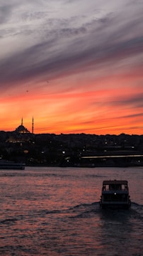A boat is sailing in the foreground, heading away from the viewer on a large body of water. The sky is painted with a stunning sunset of deep orange and red hues, contrasting against a silhouette of a city skyline with a prominent mosque featuring two tall minarets. Birds are scattered in the sky, adding life to the serene scene.
