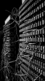 A close-up view of two stacks of old electrical components with numerous tangled wires connected to them. The image presents a complex and intricate pattern of cables that weave in and out of grid-like structures, suggesting an antiquated or retro technology setup. The black and white color scheme adds a dramatic and historical feel to the scene.