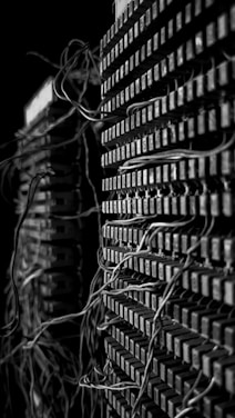 A close-up view of two stacks of old electrical components with numerous tangled wires connected to them. The image presents a complex and intricate pattern of cables that weave in and out of grid-like structures, suggesting an antiquated or retro technology setup. The black and white color scheme adds a dramatic and historical feel to the scene.