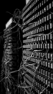 A close-up view of two stacks of old electrical components with numerous tangled wires connected to them. The image presents a complex and intricate pattern of cables that weave in and out of grid-like structures, suggesting an antiquated or retro technology setup. The black and white color scheme adds a dramatic and historical feel to the scene.