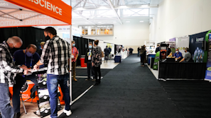 A trade show or exhibition hall with multiple booths lined along the aisles. People are interacting and exploring different displays. Some individuals are wearing masks. The hall is well-lit with banners and promotional materials displayed prominently.