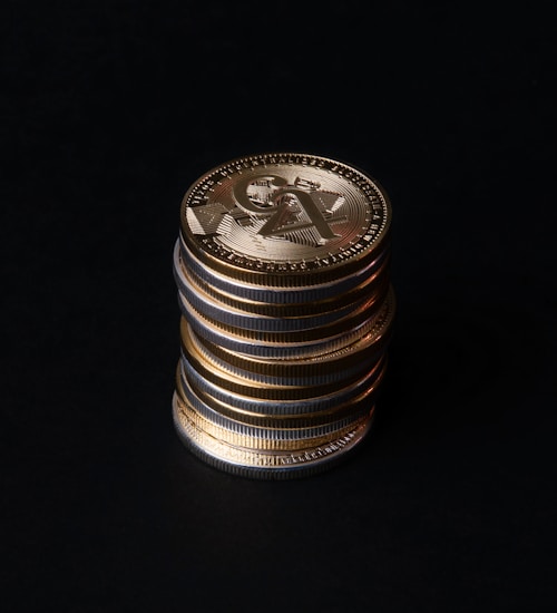 A stack of gold crypto coins with intricate patterns and symbols against a dark background, illuminated to highlight their metallic luster.