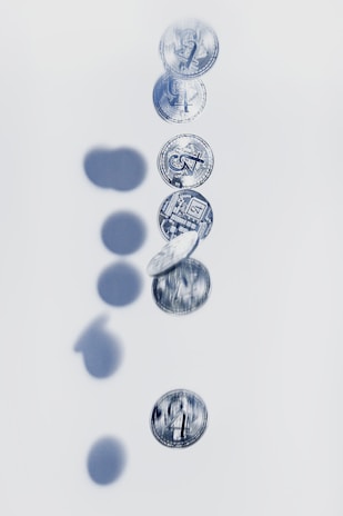 A vertical sequence of coins in motion is captured, creating a blurred effect. Shadows of the coins are visible on the light background, suggesting a dynamic fall. The coins have intricate designs and appear metallic.