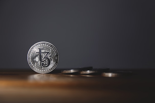A silver coin with intricate details stands upright on a wooden surface, while several other coins are laid flat in the background. The background is dark, creating a stark contrast with the shiny coins.
