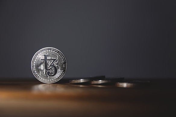 A silver coin with intricate details stands upright on a wooden surface, while several other coins are laid flat in the background. The background is dark, creating a stark contrast with the shiny coins.