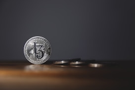 A silver coin with intricate details stands upright on a wooden surface, while several other coins are laid flat in the background. The background is dark, creating a stark contrast with the shiny coins.