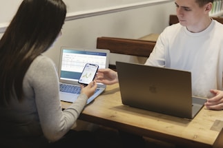 Two people are seated at a wooden table, each using a laptop. One person hands a smartphone to the other, displaying a graph or chart. The environment appears to be informal and collaborative.