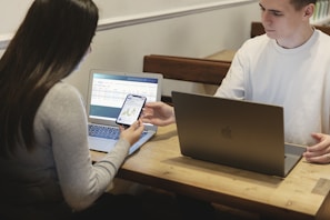 Two people are seated at a wooden table, each using a laptop. One person hands a smartphone to the other, displaying a graph or chart. The environment appears to be informal and collaborative.