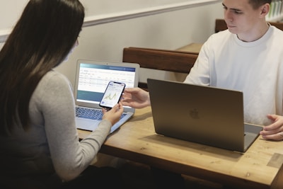 Two people are seated at a wooden table, each using a laptop. One person hands a smartphone to the other, displaying a graph or chart. The environment appears to be informal and collaborative.