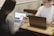 Two people are seated at a wooden table, each using a laptop. One person hands a smartphone to the other, displaying a graph or chart. The environment appears to be informal and collaborative.