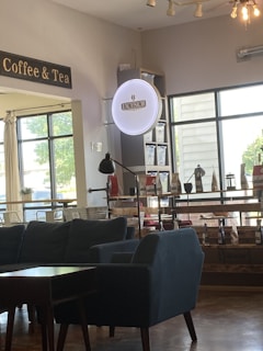 A cozy interior of a coffee shop featuring a dark blue couch and armchair set around a small table. The wall displays a sign reading 'Excelsior' next to a 'Coffee & Tea' sign. There is a bright window with natural light illuminating the space. Shelves contain various packaged coffee products, and a floor lamp adds to the warm ambiance.