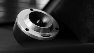 A close-up view of a metallic, circular object with a beveled edge and three screw holes. The object is set against a dark background with a partially obscured, blurred light source in the background. It has a polished finish, with a reflective surface and etched lettering.