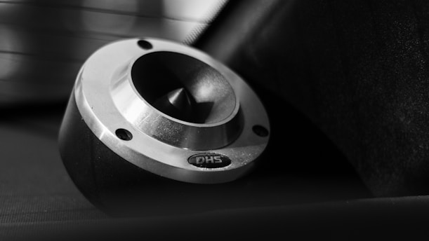 A close-up view of a metallic, circular object with a beveled edge and three screw holes. The object is set against a dark background with a partially obscured, blurred light source in the background. It has a polished finish, with a reflective surface and etched lettering.