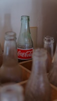 A clear glass Coca-Cola bottle with a red and white label stands among several other empty and dusty glass bottles. The background is a plain wall, and the bottles are placed in a wooden crate.