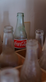 A clear glass Coca-Cola bottle with a red and white label stands among several other empty and dusty glass bottles. The background is a plain wall, and the bottles are placed in a wooden crate.