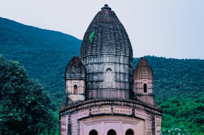 A historic temple structure with intricate architecture, featuring a large central dome flanked by two smaller domes. The building is set against a backdrop of lush green hills under a clear sky, with abundant greenery surrounding the area.
