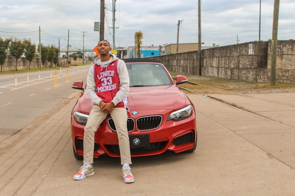 A person wearing a red and white sports jersey with the text 'Lower Merion 33' is leaning against the front of a red BMW car. The individual is also wearing beige pants, colorful sneakers, and a cap that is worn backwards. The scene is set on an urban street with concrete pavements, electric poles, and buildings in the background under a cloudy sky.