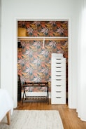 A small closet with vibrant, tropical leaf-patterned wallpaper in shades of orange, pink, and green. Inside, there is a tall, white drawer unit on the right and a black metal frame bench on the left with a wooden shelf underneath. A mustard-colored rolled blanket is placed on a shelf inside the closet.