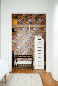 A small closet with vibrant, tropical leaf-patterned wallpaper in shades of orange, pink, and green. Inside, there is a tall, white drawer unit on the right and a black metal frame bench on the left with a wooden shelf underneath. A mustard-colored rolled blanket is placed on a shelf inside the closet.