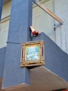A decorative framed painting is mounted on a blue wall near the top railing of a staircase. Above the painting, a red high-heeled shoe is placed on the ledge.