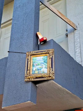 A decorative framed painting is mounted on a blue wall near the top railing of a staircase. Above the painting, a red high-heeled shoe is placed on the ledge.