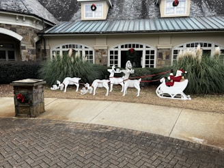 A festive holiday decoration is displayed in front of a building with stone and brickwork. It shows a white sleigh with a figure in red, resembling Santa Claus, being pulled by four reindeer cutouts. The setup is arranged on a small patch of grass, with large windows and some ornamental grass behind it. Red bows are featured on a nearby stone post and on the building's windows.