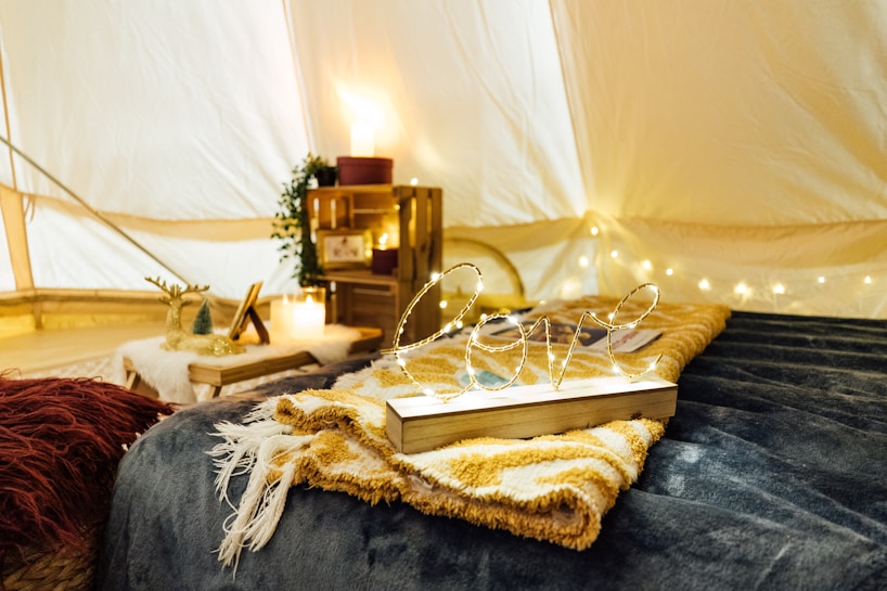A cozy interior scene with a warm, intimate atmosphere. A wooden sign with the word 'love' scripted in fairy lights is placed on a folded blanket. The background features a small table adorned with candles and decorative items, including miniature trees and a photo frame. Soft lighting creates a serene ambiance, enhanced by string lights along the tent's fabric walls.