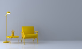 A minimalist interior setting features a bright yellow armchair, table, and lamp against a neutral gray wall. A small plant in a matching yellow pot sits on the table, adding a touch of greenery.