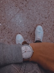 A person wearing a gray sweater, looking down at their wristwatch with a brown leather strap and a white face displaying the brand name and the date. The background features a speckled textured floor, and the person's white sneakers are partially visible at the bottom of the image.