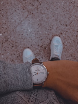 A person wearing a gray sweater, looking down at their wristwatch with a brown leather strap and a white face displaying the brand name and the date. The background features a speckled textured floor, and the person's white sneakers are partially visible at the bottom of the image.