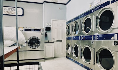 A laundry room featuring a series of large industrial washing and drying machines along a wall. The machines are metallic with blue trim and digital displays, suggesting they are coin-operated. A door is visible in the background. In the foreground, there is a metal laundry basket and a white cloth on a folding counter.