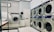 A laundry room featuring a series of large industrial washing and drying machines along a wall. The machines are metallic with blue trim and digital displays, suggesting they are coin-operated. A door is visible in the background. In the foreground, there is a metal laundry basket and a white cloth on a folding counter.