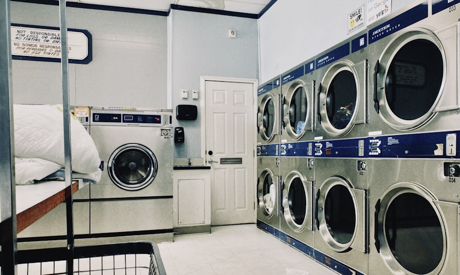 A laundry room featuring a series of large industrial washing and drying machines along a wall. The machines are metallic with blue trim and digital displays, suggesting they are coin-operated. A door is visible in the background. In the foreground, there is a metal laundry basket and a white cloth on a folding counter.