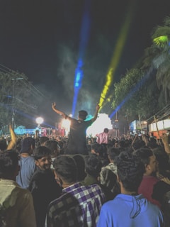A large crowd of people gathered at an outdoor event during nighttime, with colorful stage lights illuminating the scene. A person is raised above the crowd, silhouetted against bright blue and yellow beams of light. The atmosphere is lively and energetic, with trees and power lines visible in the background.