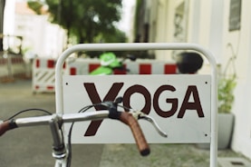 A sign with the word 'YOGA' in brown letters is affixed to a metal frame. A bicycle handlebar with brown grips is in the foreground, partially blocking the sign. In the background, there are blurred details of a street with construction barriers and trees.