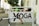 A sign with the word 'YOGA' in brown letters is affixed to a metal frame. A bicycle handlebar with brown grips is in the foreground, partially blocking the sign. In the background, there are blurred details of a street with construction barriers and trees.