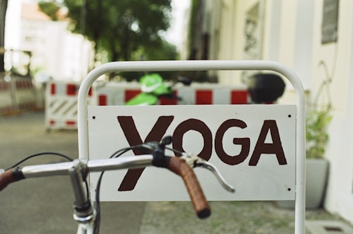 A sign with the word 'YOGA' in brown letters is affixed to a metal frame. A bicycle handlebar with brown grips is in the foreground, partially blocking the sign. In the background, there are blurred details of a street with construction barriers and trees.