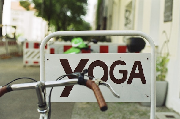 A sign with the word 'YOGA' in brown letters is affixed to a metal frame. A bicycle handlebar with brown grips is in the foreground, partially blocking the sign. In the background, there are blurred details of a street with construction barriers and trees.