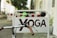 A sign with the word 'YOGA' in brown letters is affixed to a metal frame. A bicycle handlebar with brown grips is in the foreground, partially blocking the sign. In the background, there are blurred details of a street with construction barriers and trees.