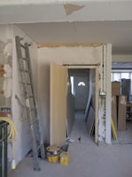 The image depicts a partially renovated or under-construction room. An unfinished wall with visible drywall and joints can be seen, alongside scattered construction materials like a ladder, wooden boards, and yellow construction buckets. A door is propped open, leading to a dimly lit area with more materials stacked in the background.