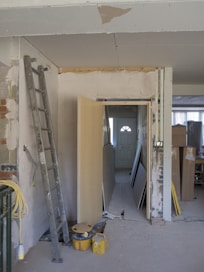 The image depicts a partially renovated or under-construction room. An unfinished wall with visible drywall and joints can be seen, alongside scattered construction materials like a ladder, wooden boards, and yellow construction buckets. A door is propped open, leading to a dimly lit area with more materials stacked in the background.