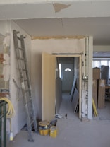 The image depicts a partially renovated or under-construction room. An unfinished wall with visible drywall and joints can be seen, alongside scattered construction materials like a ladder, wooden boards, and yellow construction buckets. A door is propped open, leading to a dimly lit area with more materials stacked in the background.