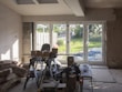 A room under renovation with construction materials scattered on the floor. There's a large window letting in sunlight and providing a view of a lush garden outside. Various tools and boxes are on a workbench, and insulation material is piled on the left.